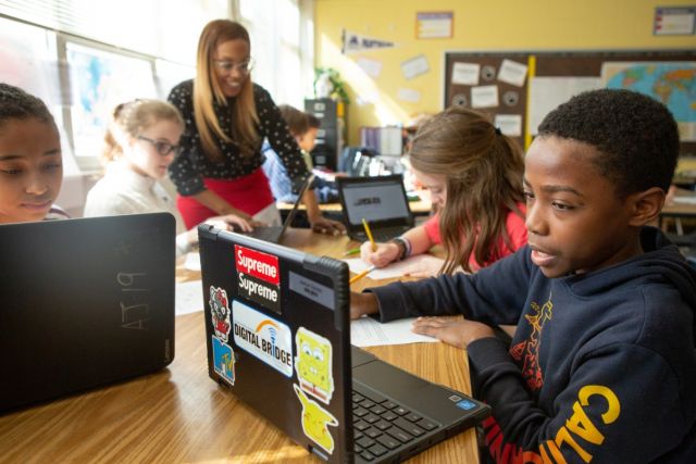 kids working on laptops with teacher in background